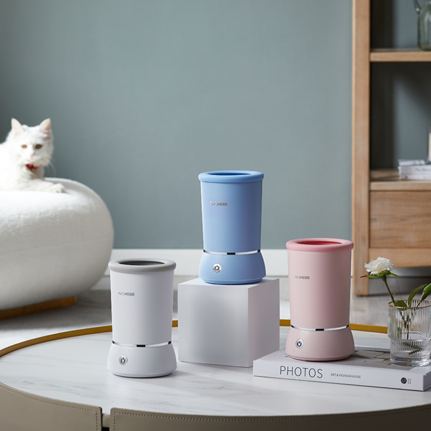 Three small cylindrical humidifiers in white, blue, and pink on a table with a cat in the background.