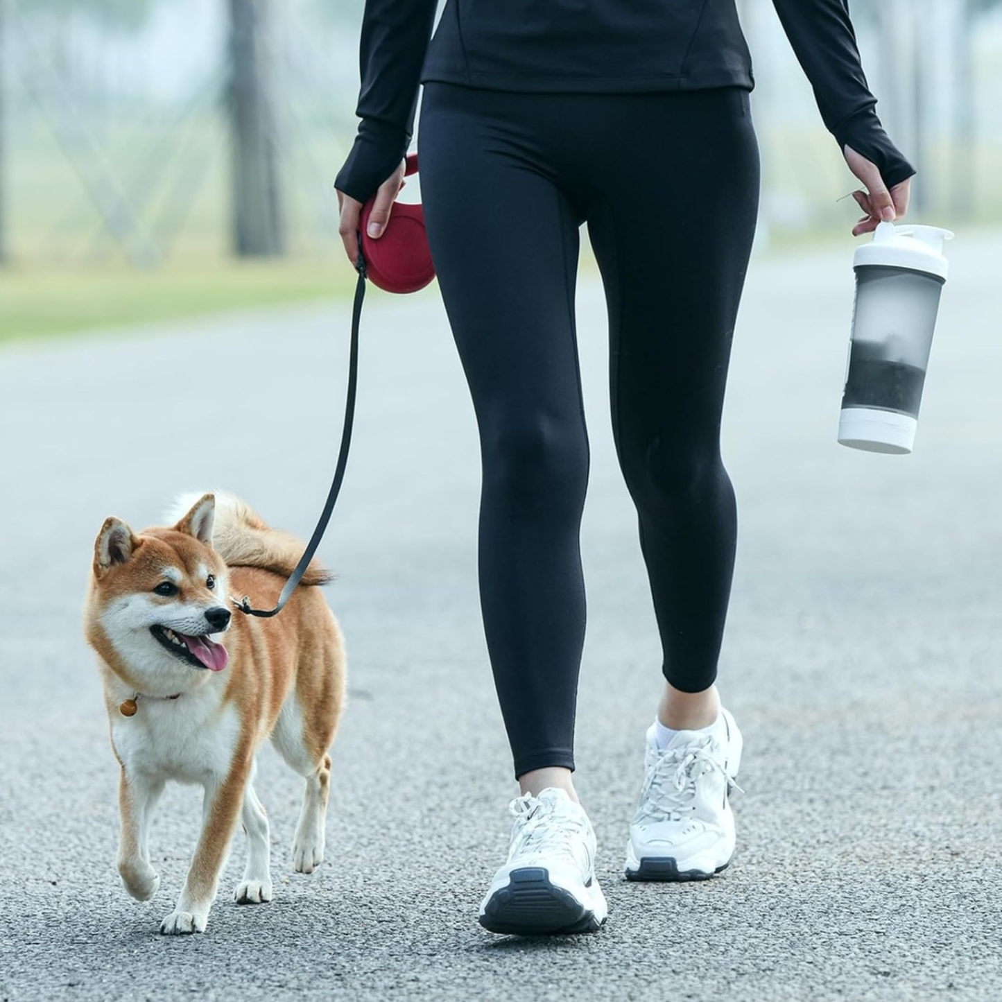 Person walking a dog on a leash with a cup in hand, wearing black leggings and white sneakers.