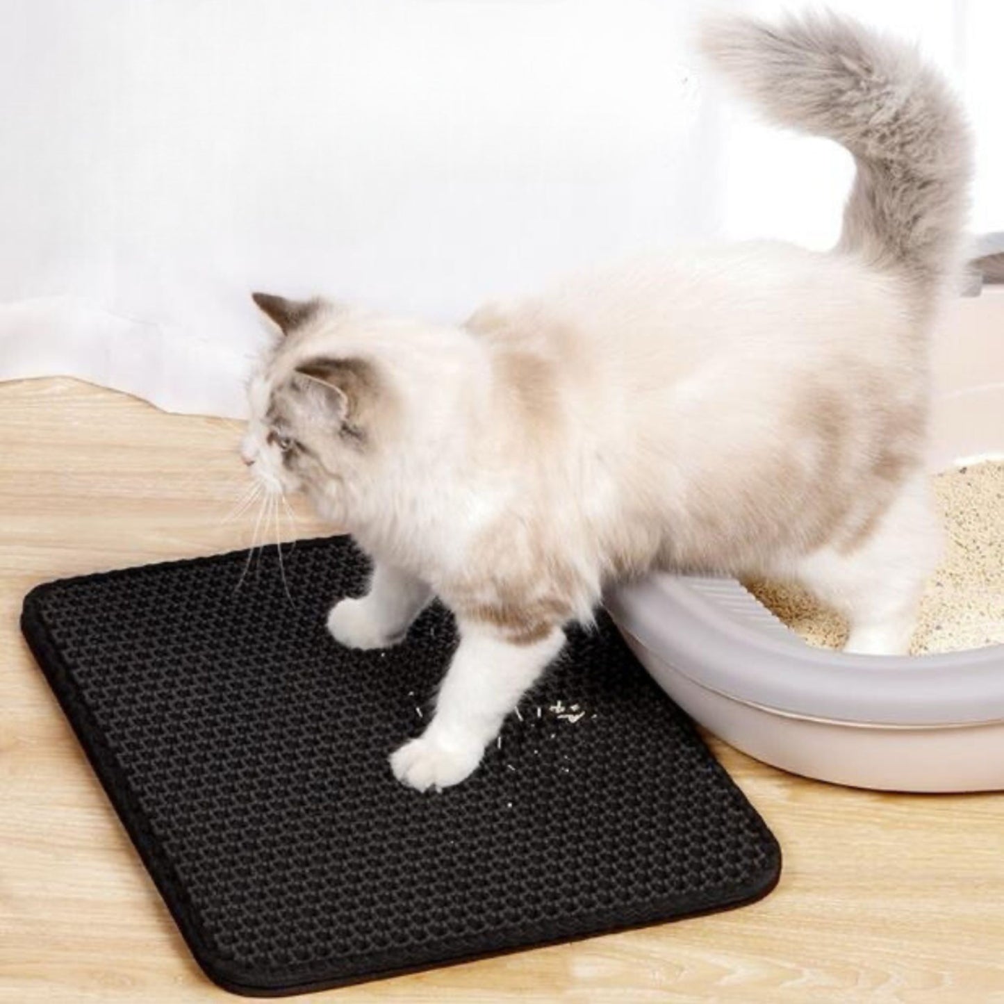 Cat using a black litter mat next to a litter box on a wooden floor.